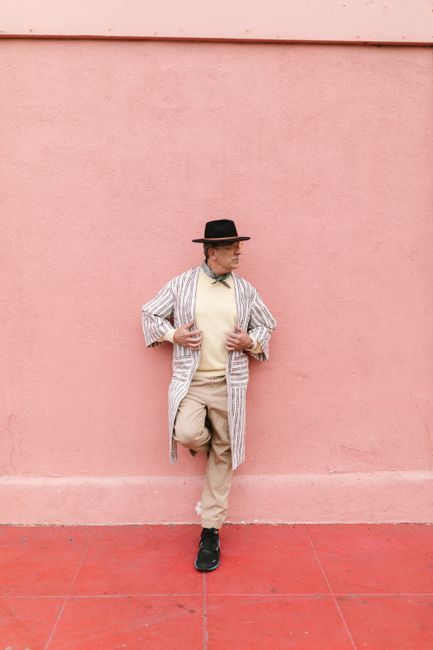 Elderly man in fashionable attire posing confidently against a vibrant pink wall outdoors.