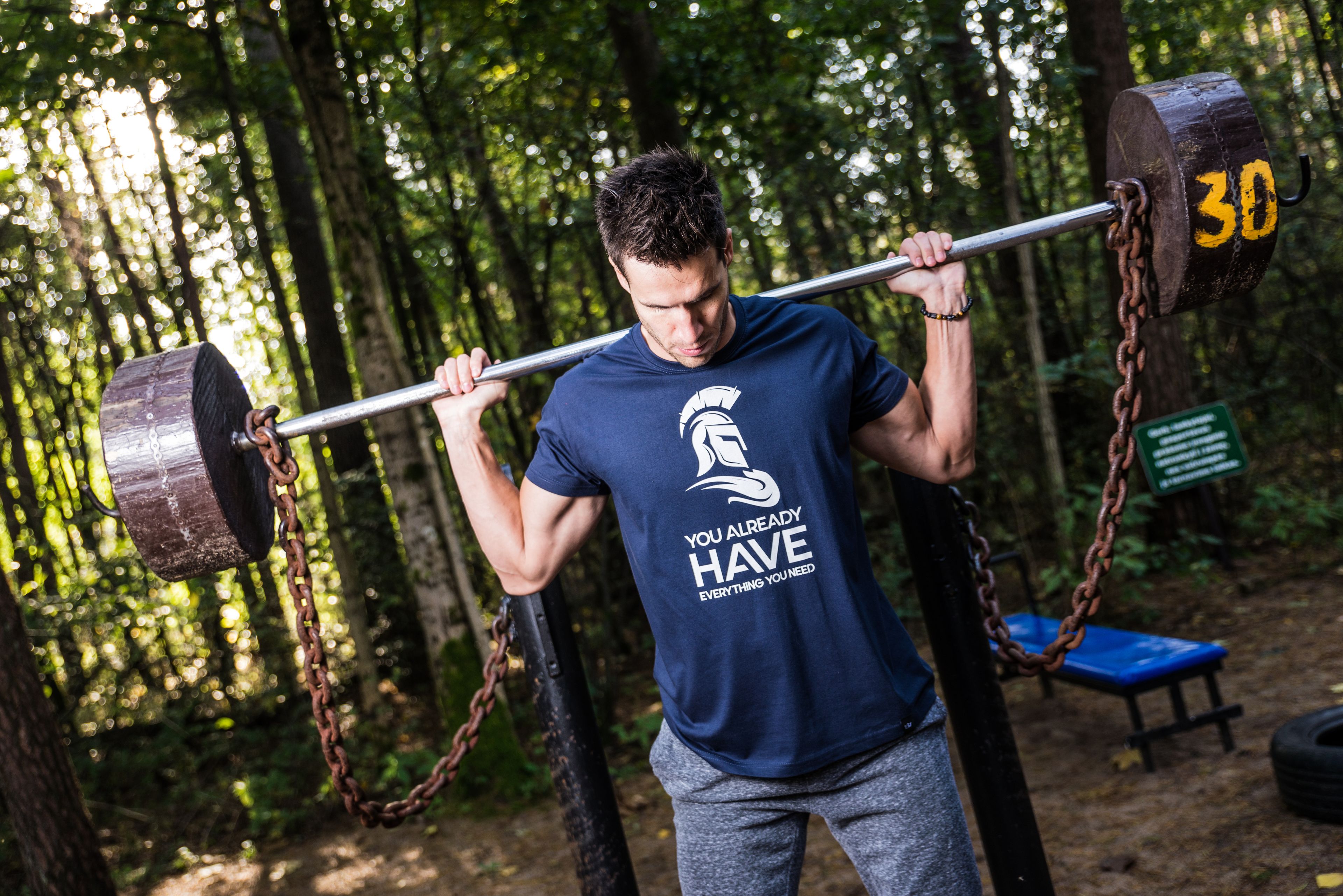Man lifting barbell with chains in a wooded outdoor gym, showcasing strength training.