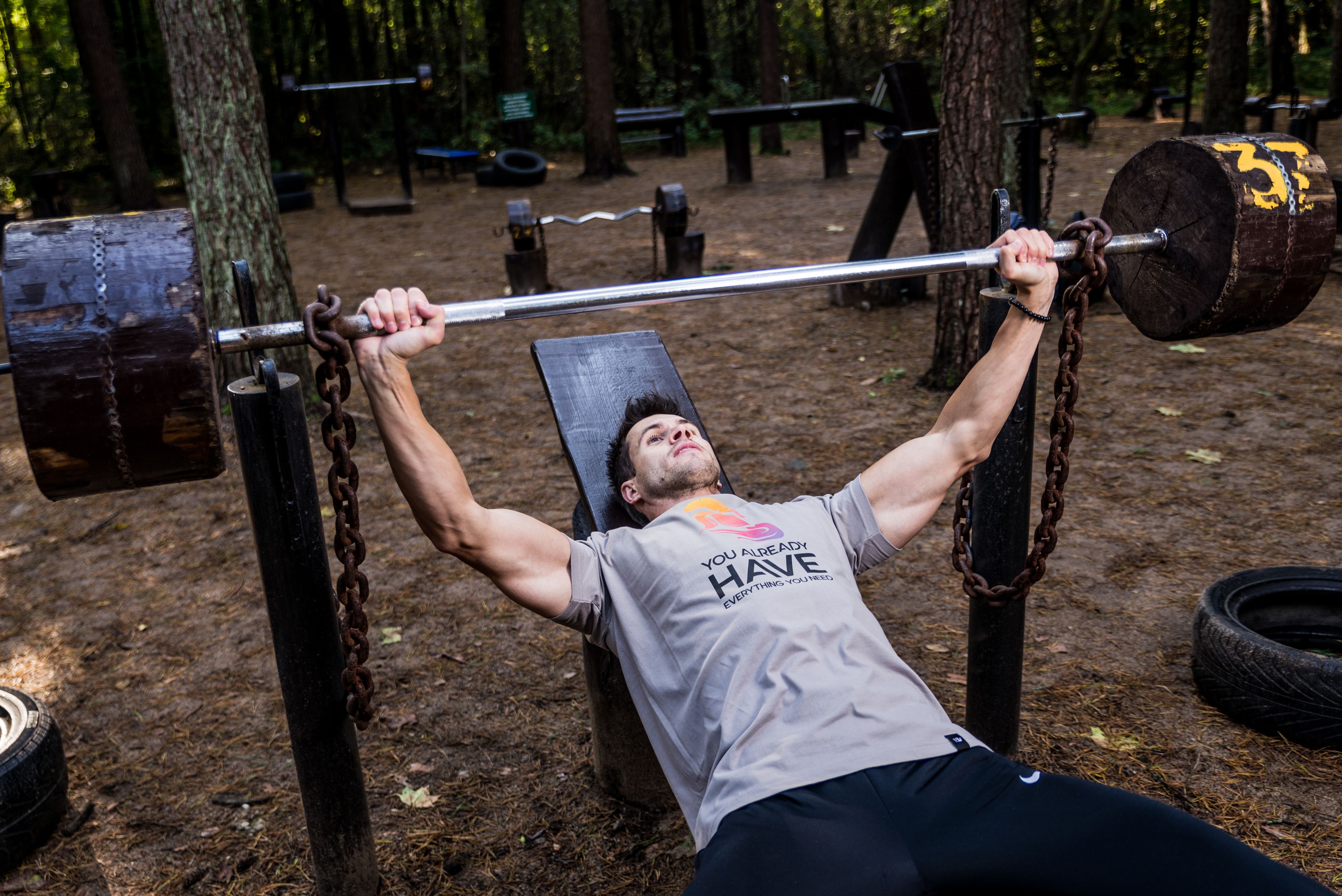 Man weightlifting outdoors, using a bench press setup amid trees for strength training.