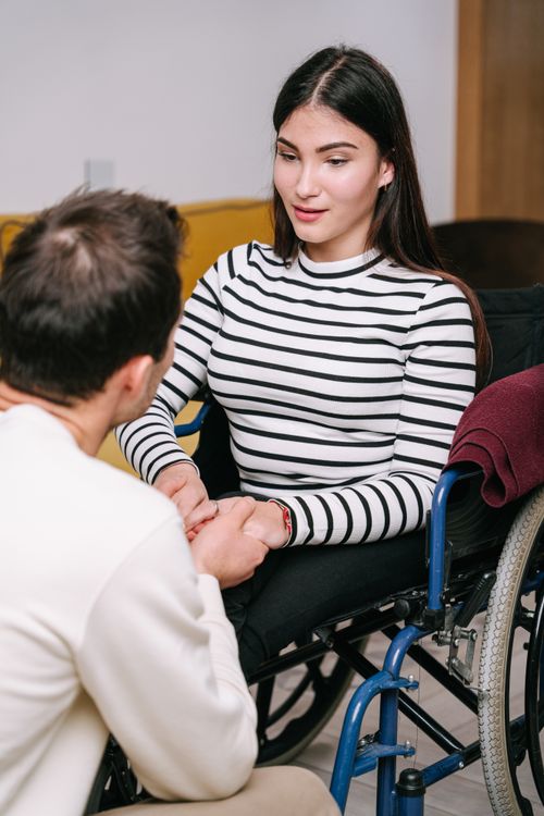 Free Young couple sitting together, holding hands, with one person in a wheelchair, expressing care. Stock Photo