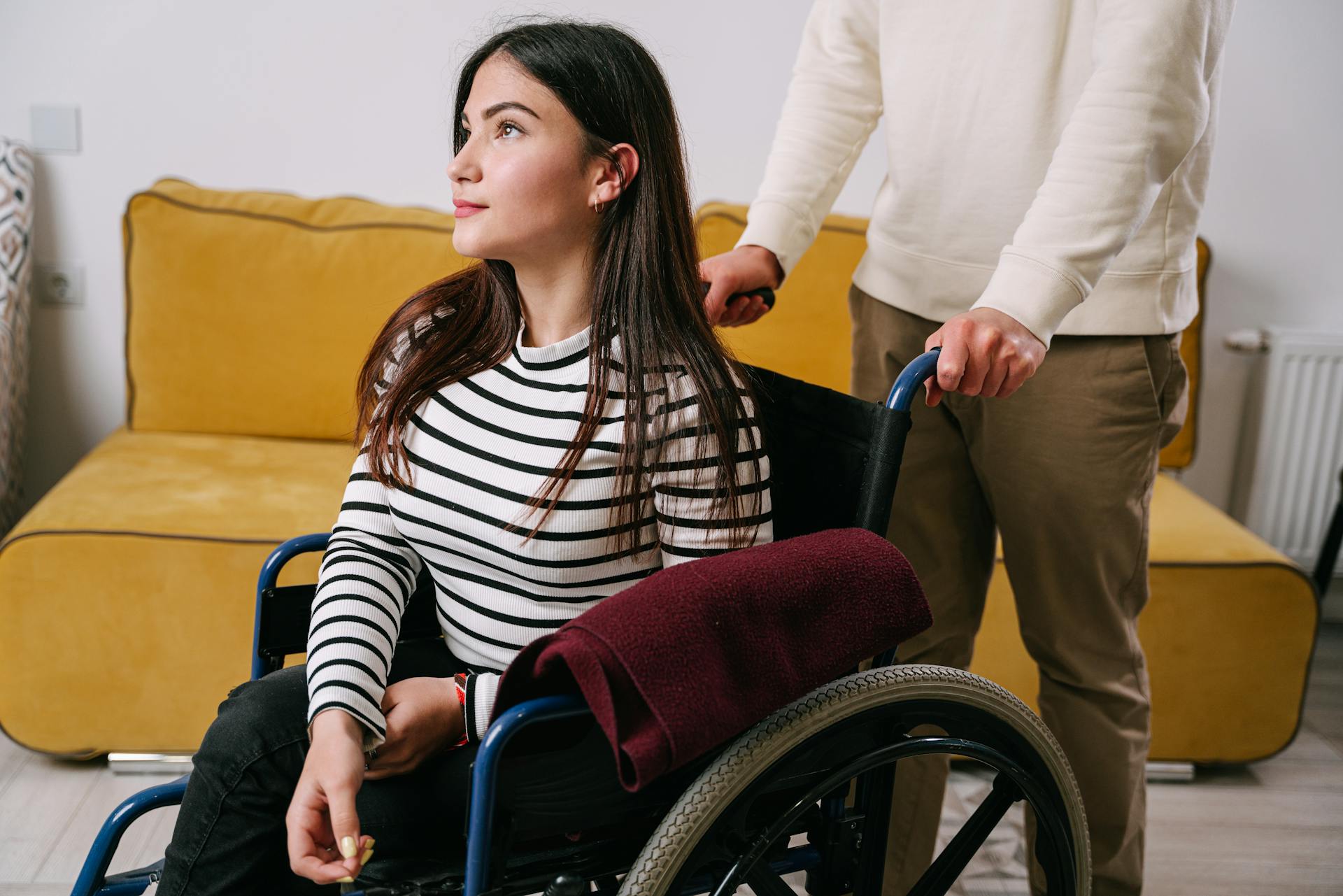 A young woman in a wheelchair being assisted indoors by a caregiver, showcasing support and mobility.