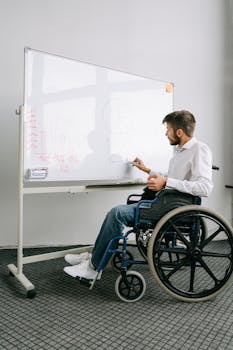 Man in a wheelchair writing on a whiteboard, presenting ideas in an office setting.