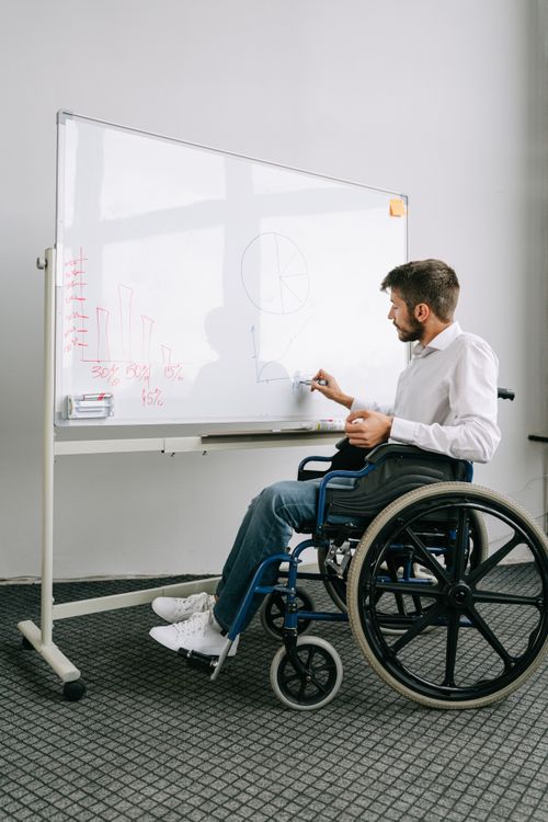 Free Man in a wheelchair writing on a whiteboard, presenting ideas in an office setting. Stock Photo