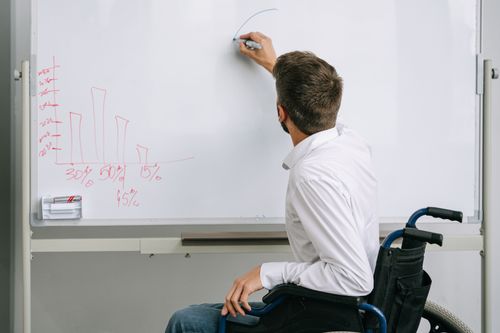 Free Man in a wheelchair writing on a whiteboard during an office presentation. Inclusive workplace environment. Stock Photo