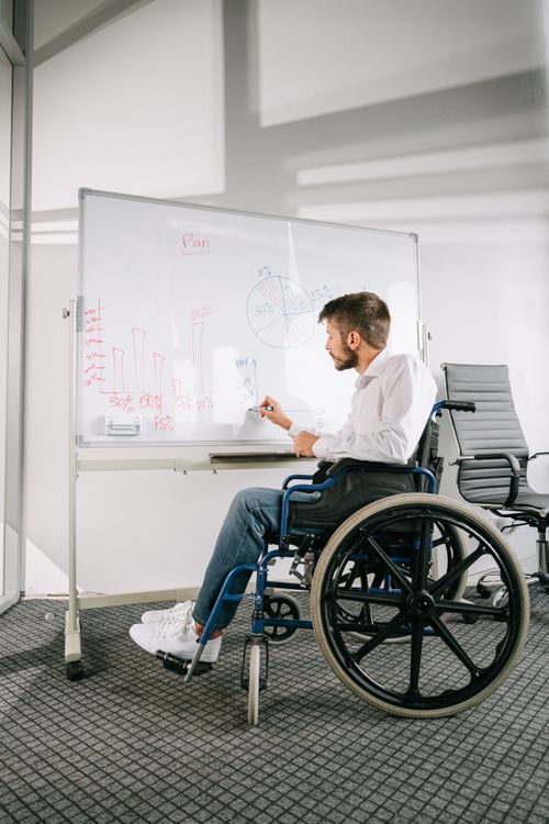 Free A young man in a wheelchair works at a whiteboard in an office setting, highlighting disability inclusion. Stock Photo