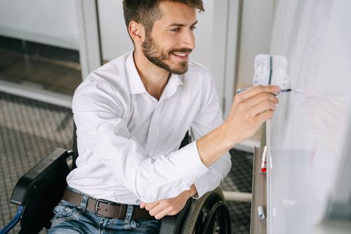Free A cheerful man in a wheelchair writing ideas on a whiteboard indoors. Stock Photo
