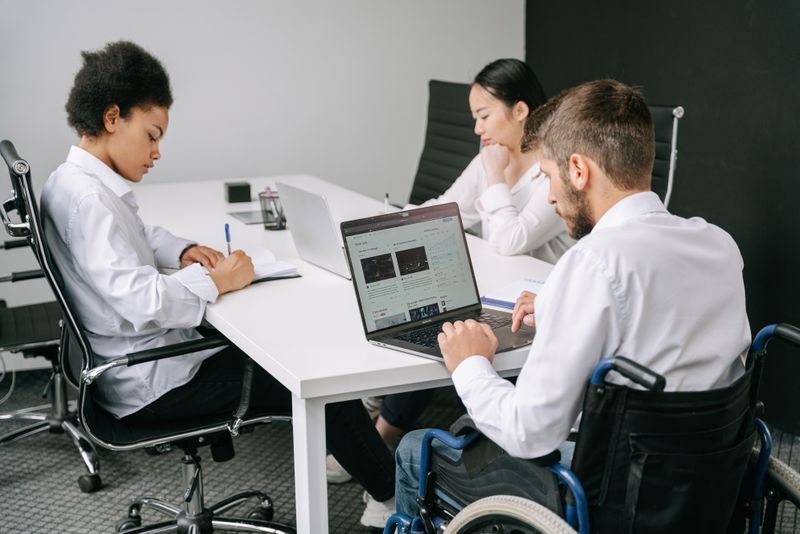 A diverse group of people with different disabilities collaborating around a table, using accessible tools and devices.