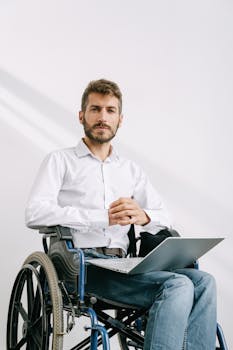 A bearded man in a wheelchair working on a laptop indoors against a white background.