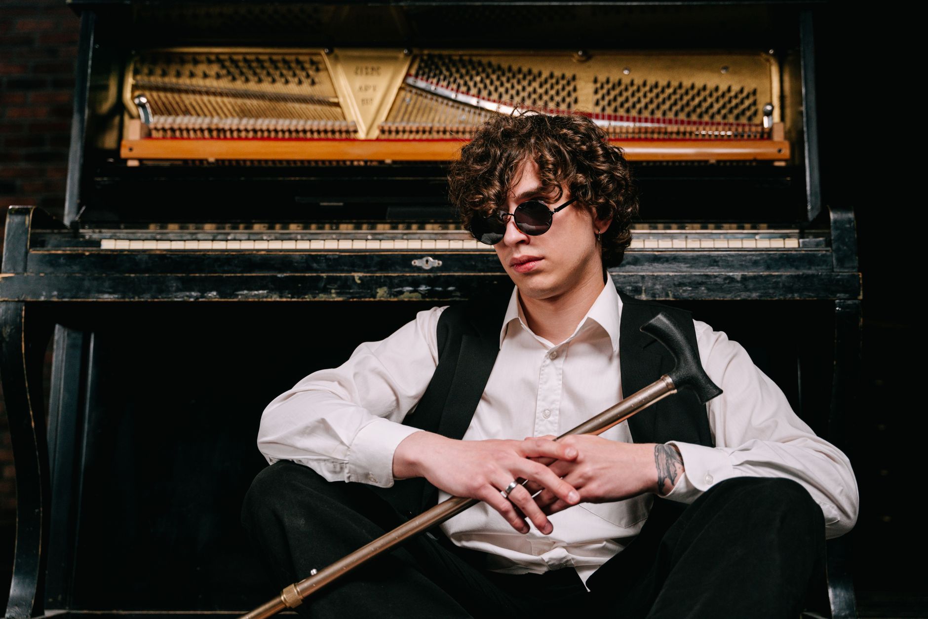 Blind musician in a white shirt and black vest sits by piano holding a walking stick.