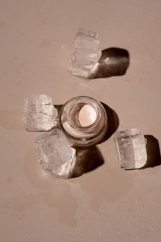 Flat lay of a skincare product surrounded by ice cubes on a textured surface.