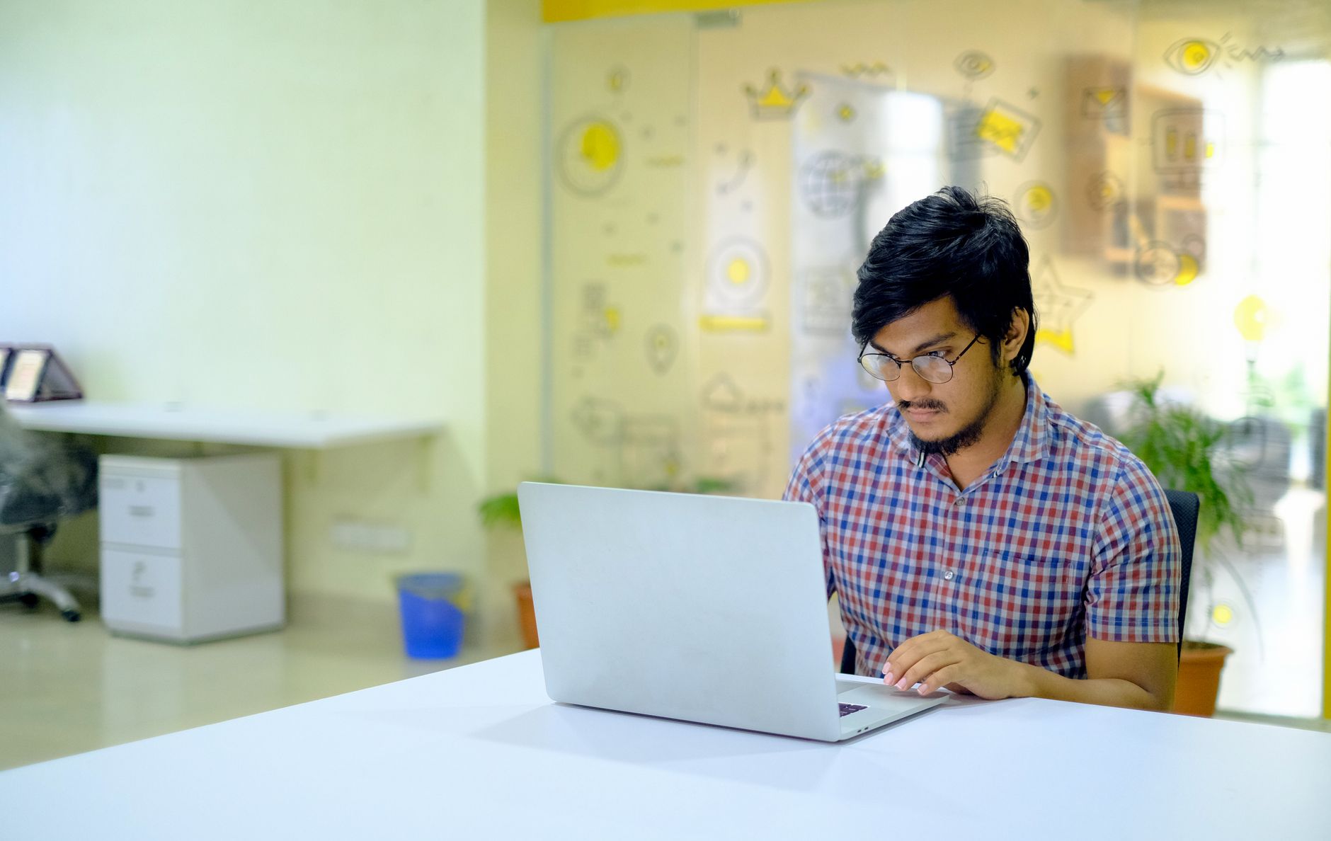 serp checker - Asian man wearing glasses works on laptop in a modern, bright office space.