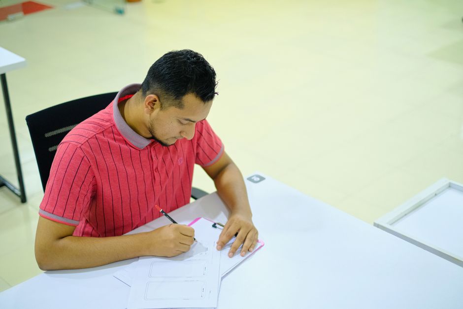 Man wearing a pink polo shirt concentrating while writing at a desk indoors.