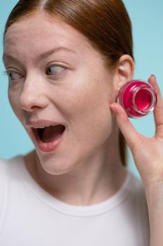 Excited woman holding a jar of bright pink cosmetic cream against a blue background.