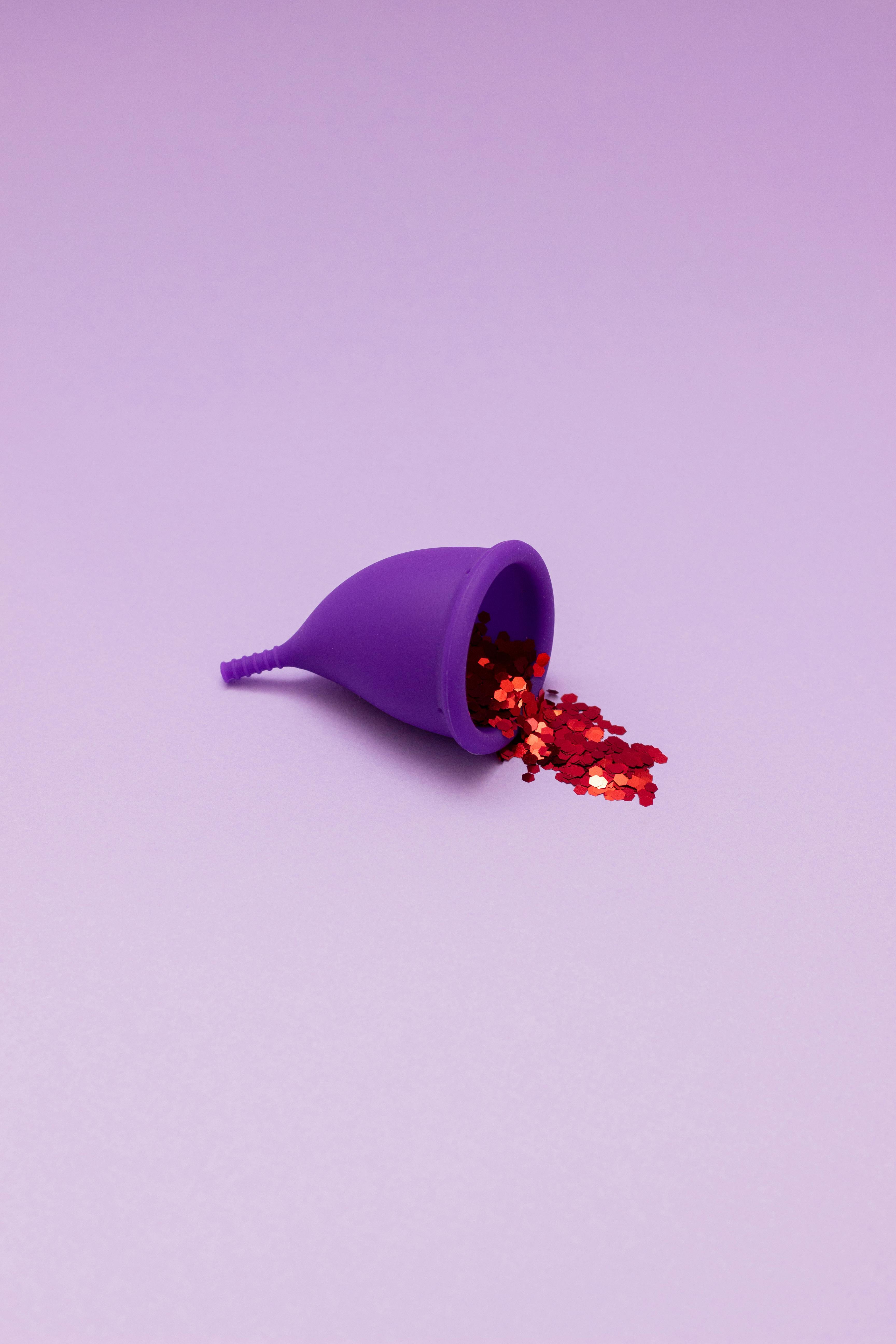 A reusable purple menstrual cup filled with red glitter, set against a lavender background, highlighting eco-friendly feminine hygiene.