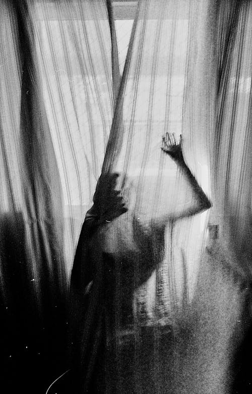 Grayscale Photo of Woman Behind the Curtains