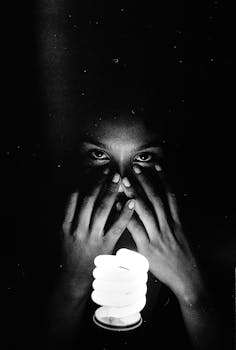 Dramatic black and white portrait with a focus on hands covering a face, illuminated by a light bulb.