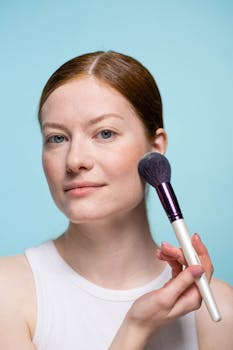 Close-up of a woman applying blush with a makeup brush against a blue background.