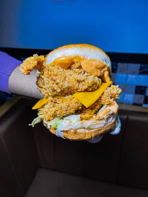 Free Close-up of a crispy chicken burger with cheddar, held in hand, showcasing delicious layers. Stock Photo
