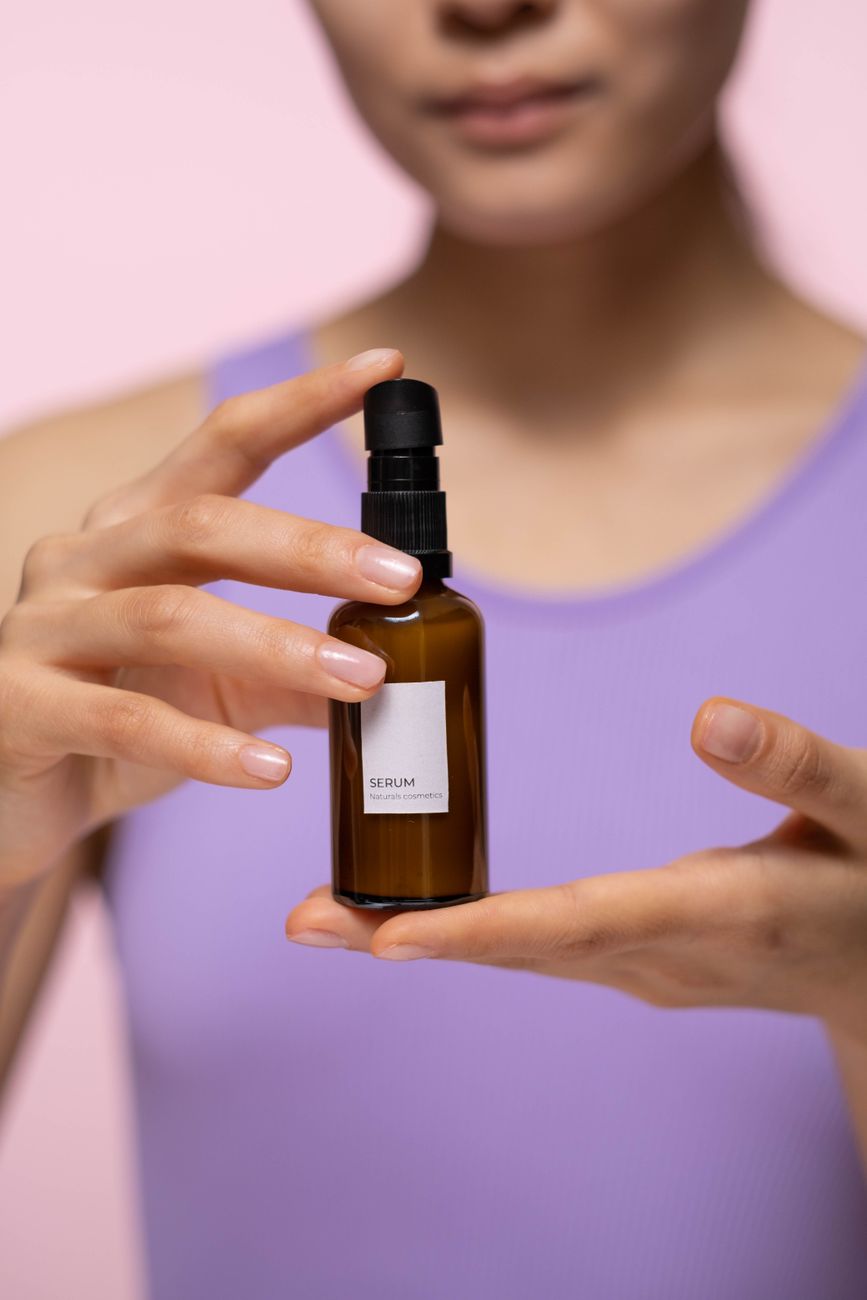 Close-up of a woman holding a skincare serum bottle indoors with a pink background.