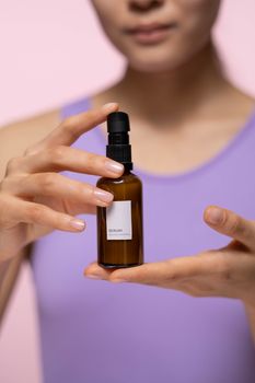 Close-up of a woman holding a skincare serum bottle indoors with a pink background.