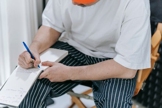 A man sits indoors writing notes with a pen in a notepad. Casual setting.