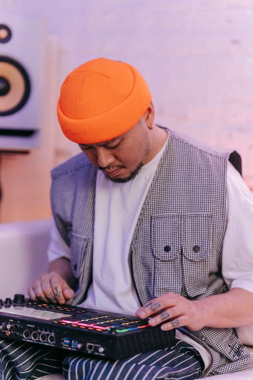 Free Asian musician in orange beanie using synth and drum machine in cozy studio setting. Stock Photo