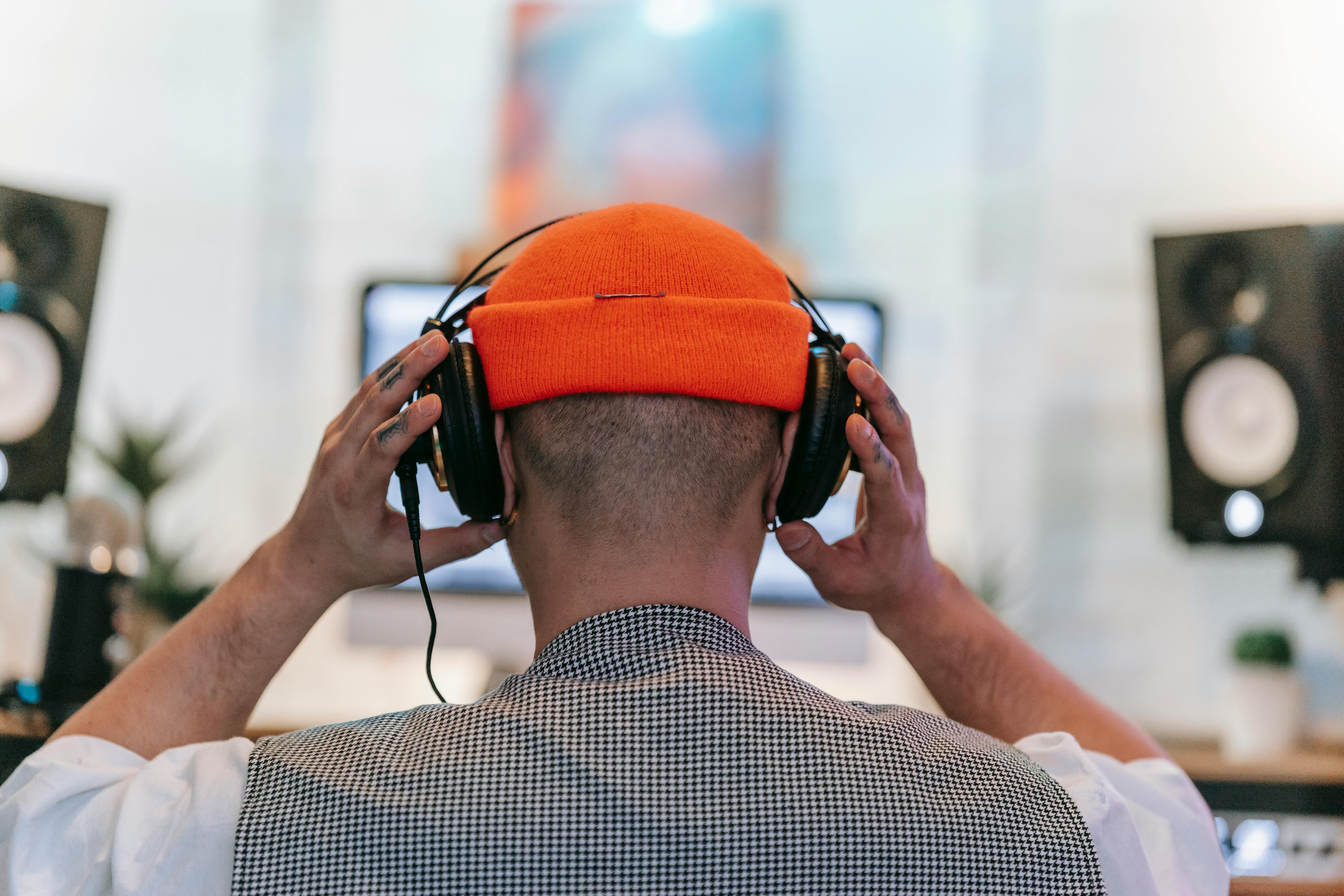 Person wearing headphones and orange beanie, focused on music production in a studio setting.