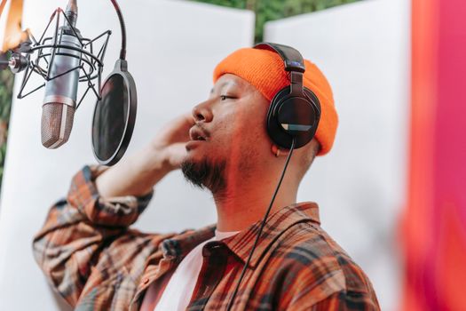 Asian man singing in a music studio wearing headphones and orange beanie with a microphone.