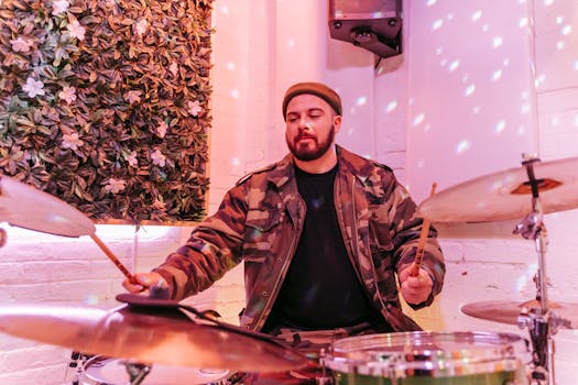 A drummer in a camouflage jacket performing indoors next to floral decor, immersed in music.