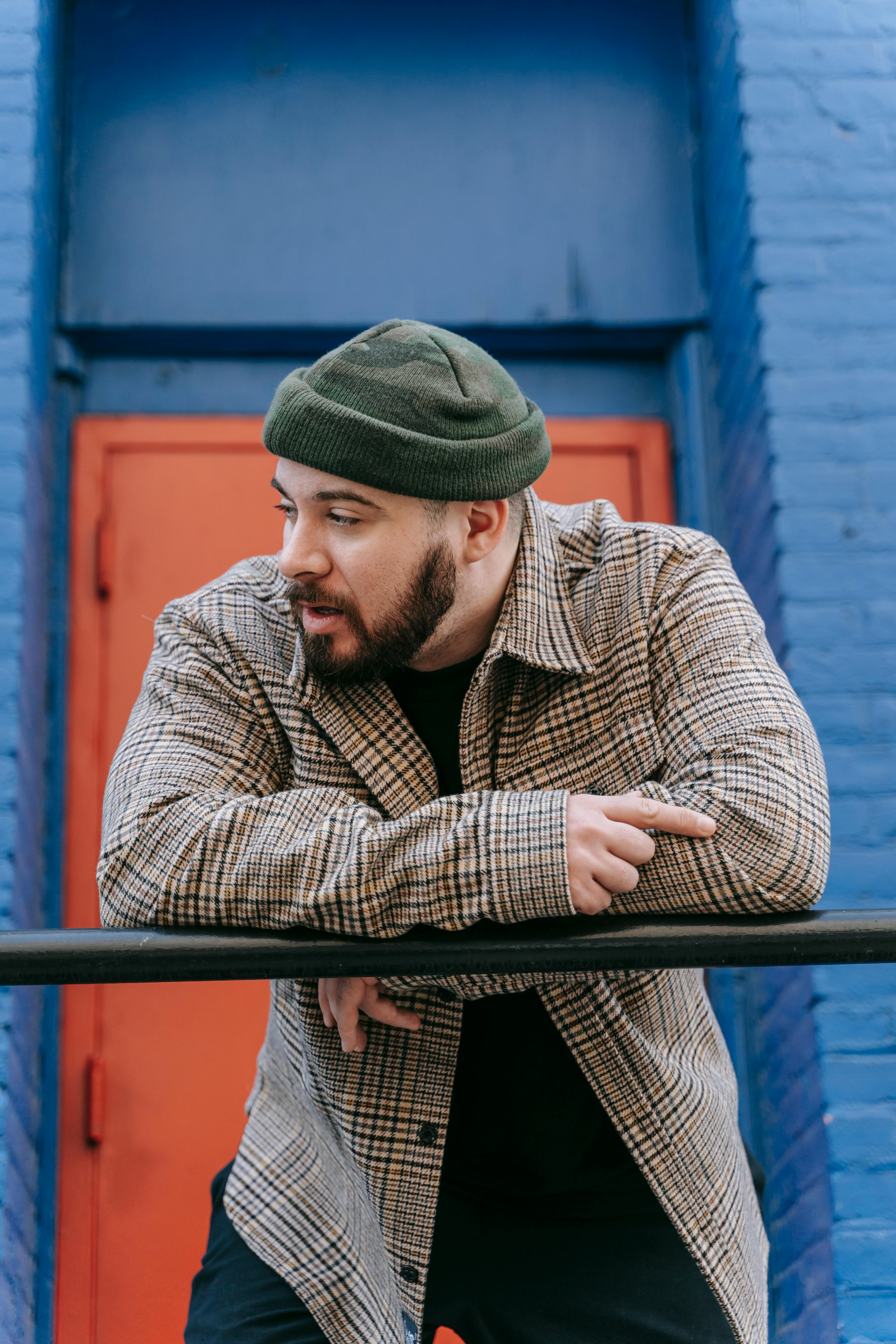 Stylish bearded man in plaid shirt and beanie leaning on a railing with colorful urban background.