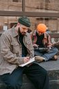 Man in Brown  Plaid Dress Shirt Writing on Book Beside a Man Playing Music