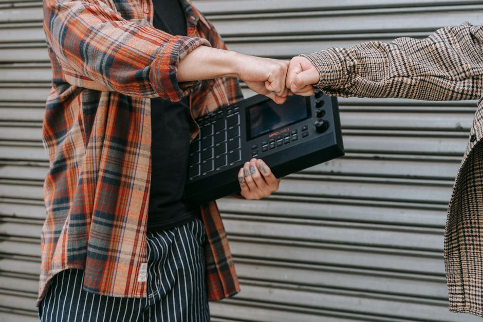 Two people in plaid shirts exchanging a fist bump while holding music equipment against a metal backdrop. Two people in plaid shirts exchanging a fist bump while holding music equipment against a metal backdrop.