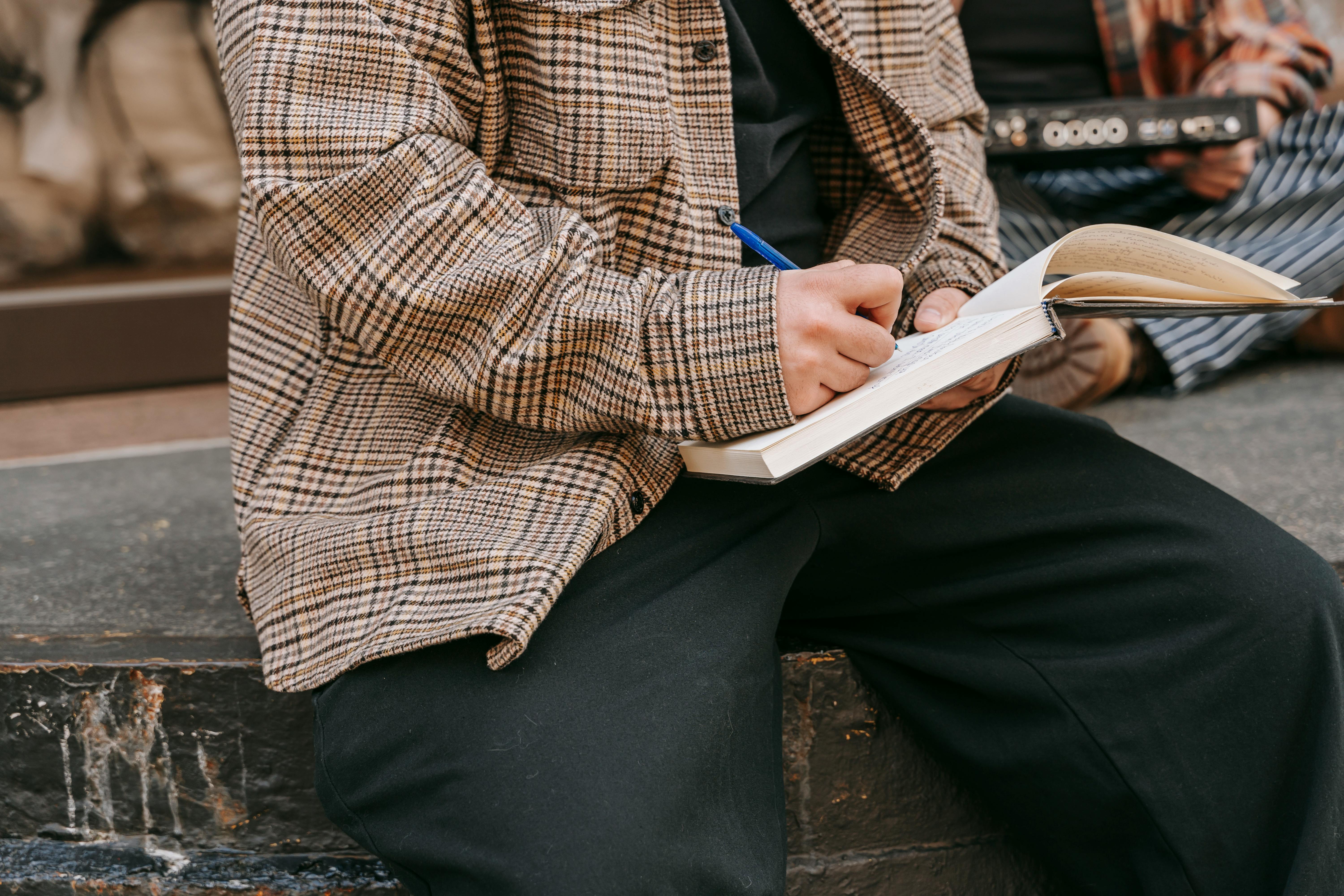Close-up of a person writing in a notebook with a pen, sitting outdoors on a sunny day.