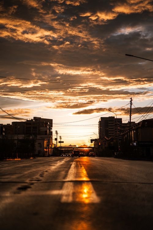 Free Silhouetted buildings line a street under dramatic sunset skies, casting reflections across the road. Stock Photo