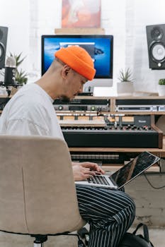 Man using laptop in music studio, side view, wearing orange beanie and casual attire.
