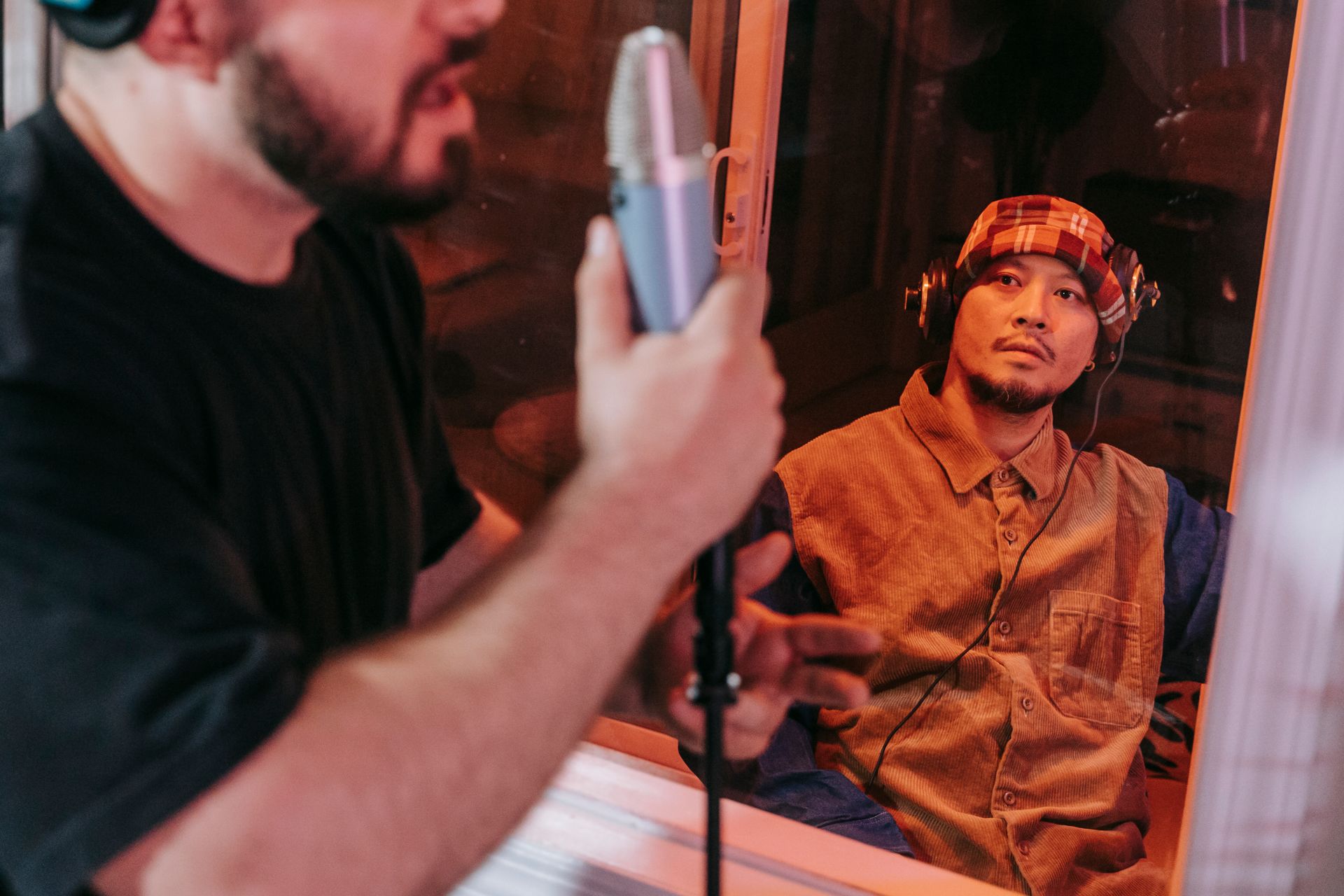 Asian musician in headphones guides a recording session from behind studio glass.
