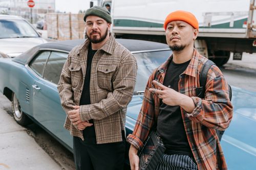 Free Two men in beanies pose confidently with a vintage car on an urban street, exuding style. Stock Photo