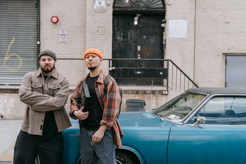 Free Two men pose confidently beside a classic car in an urban setting, showcasing streetwear fashion. Stock Photo