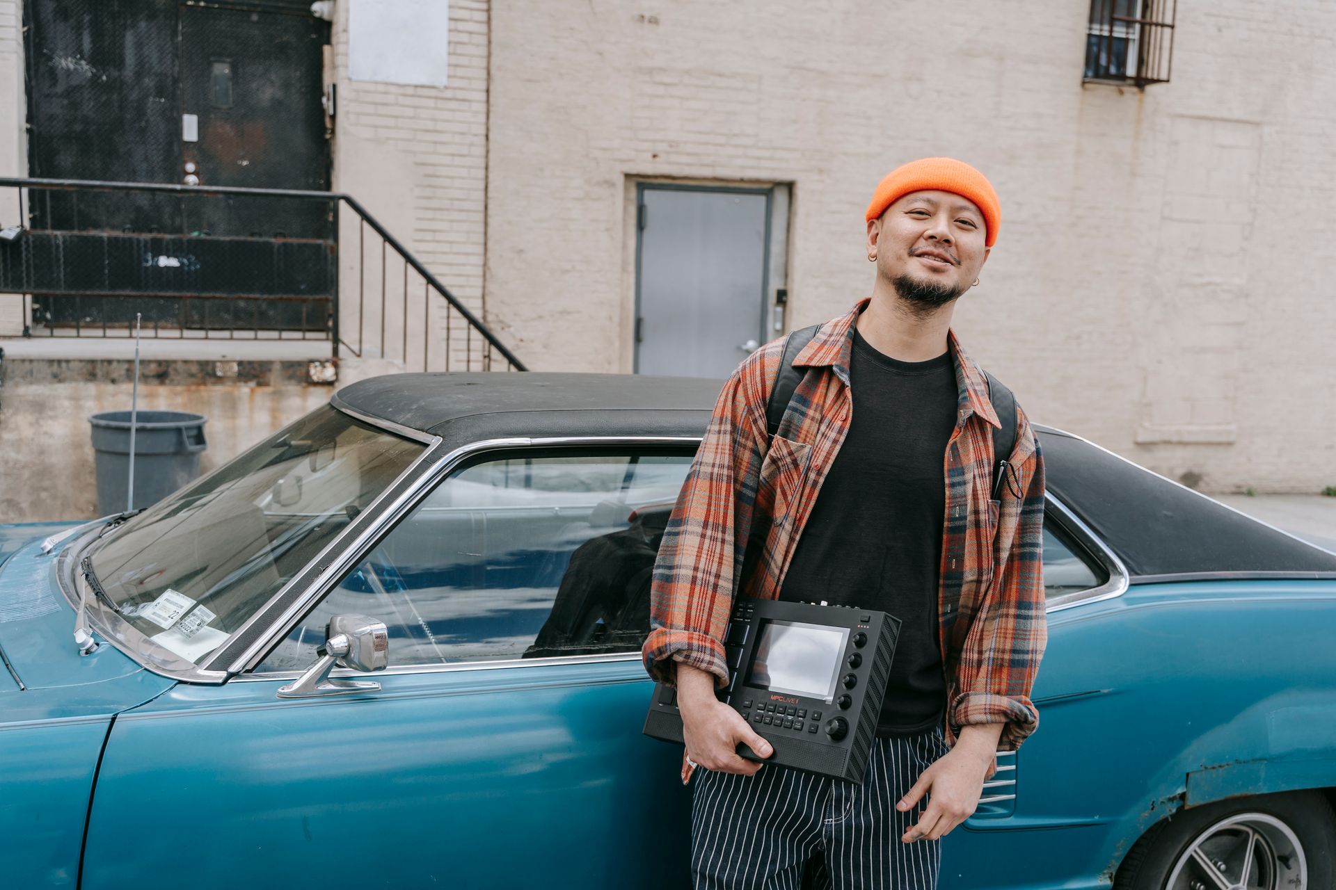 A stylish man in a knit cap leans on a vintage car holding an electronic device outdoors.