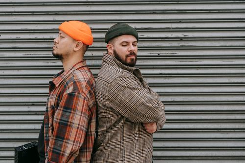 Free Two men in stylish attire stand back to back in front of a metal shutter, showcasing fashion and attitude. Stock Photo