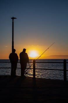 Two fishermen silhouetted against a stunning sunset over the ocean, casting their lines from a pier.