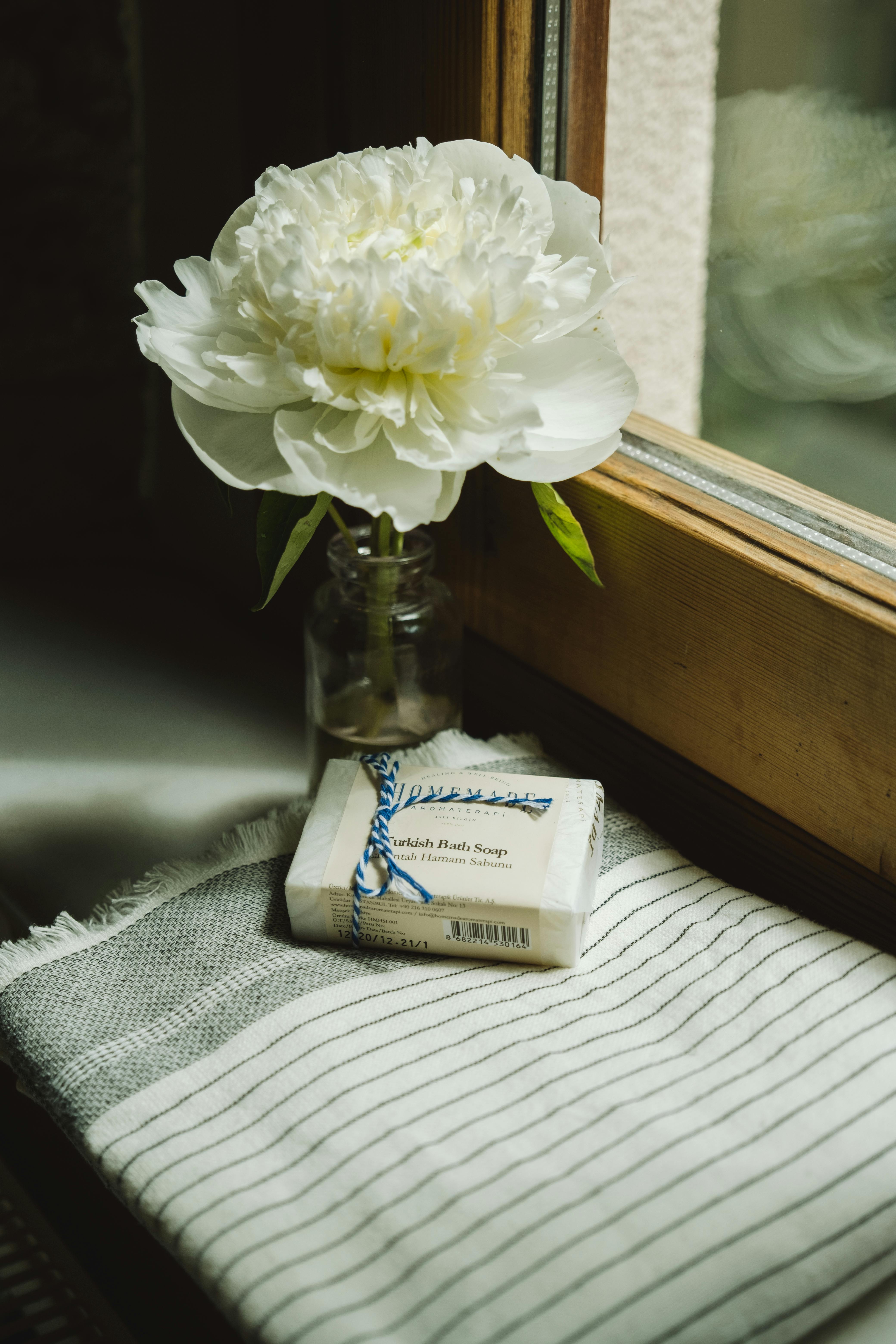 A serene still life of a white peony in a vase next to artisanal soap on a striped cloth by a window.
