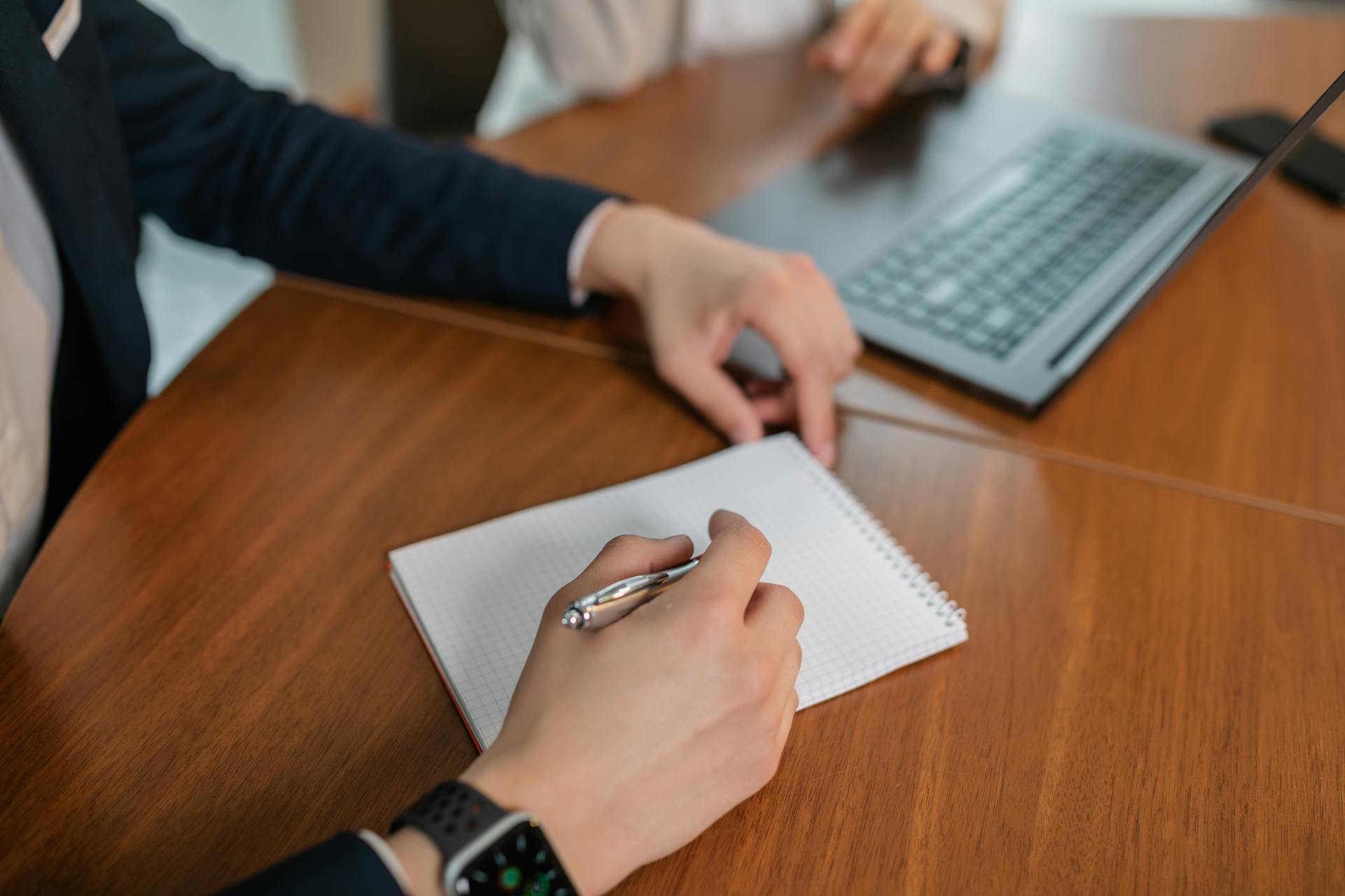 Close-up of hands writing notes on paper during a business meeting indoors with a laptop on a wooden table.