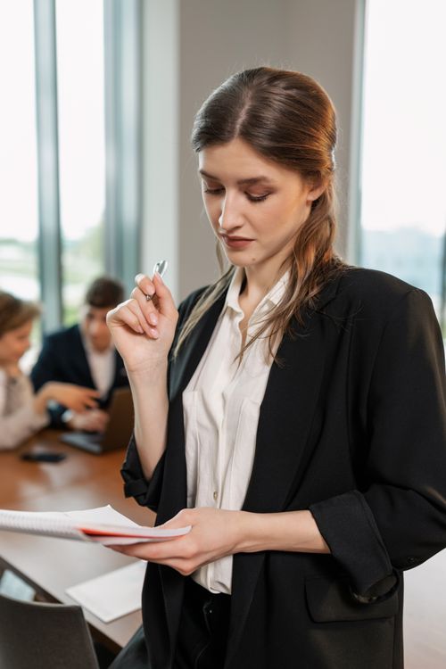 Free Businesswoman in a meeting holding documents and pen while colleagues collaborate. Stock Photo