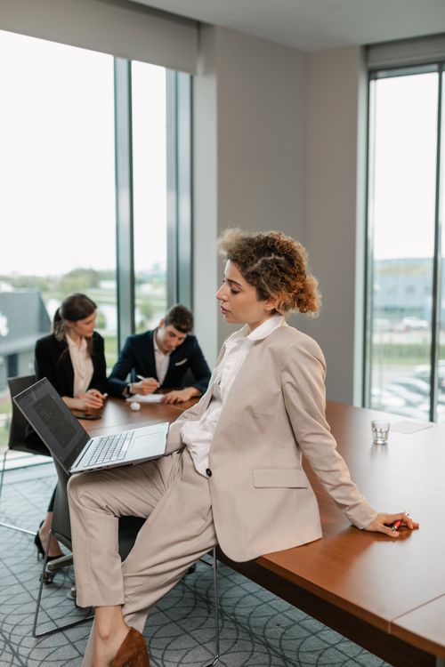 Free A woman in a beige blazer working on a laptop in a modern office setting with colleagues. Stock Photo
