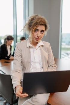 Confident businesswoman using laptop at office desk with coworkers in the background.