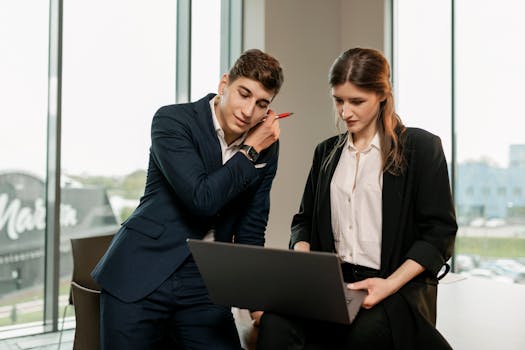 Two professionals in office attire collaborating on a laptop near large windows, enhancing teamwork and productivity.
