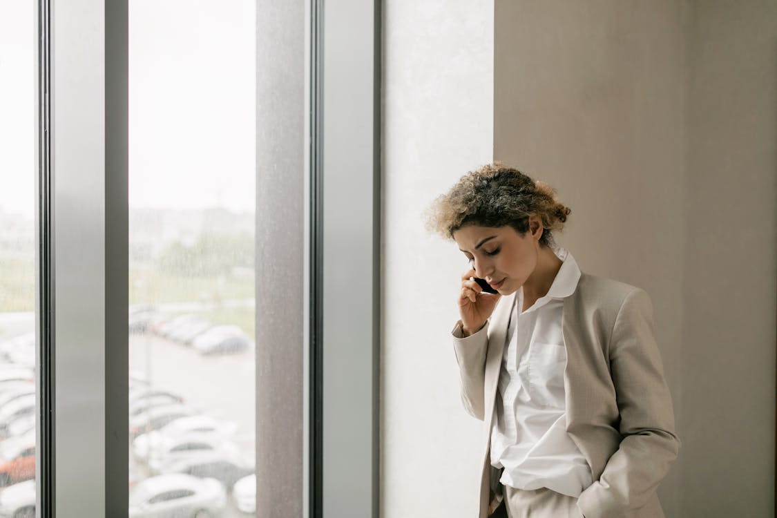 Free A Woman Standing Beside Window while Talking on the Phone Stock Photo