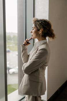 Professional woman in suit looking thoughtfully out a glass window, suggesting corporate environment.