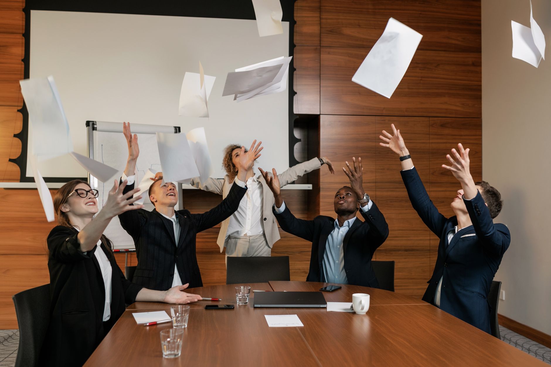 A diverse group of professionals celebrating victory in an office meeting by throwing papers in the air.